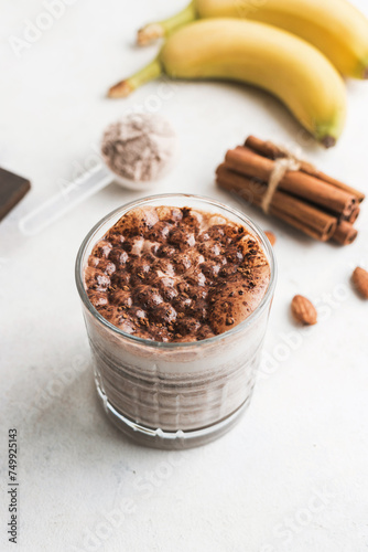 Glass with chocolate protein drink, milkshake smoothie on white table with bananas, protein powder in measuring spoon, protein bar, almond nuts and cinnamon sticks
