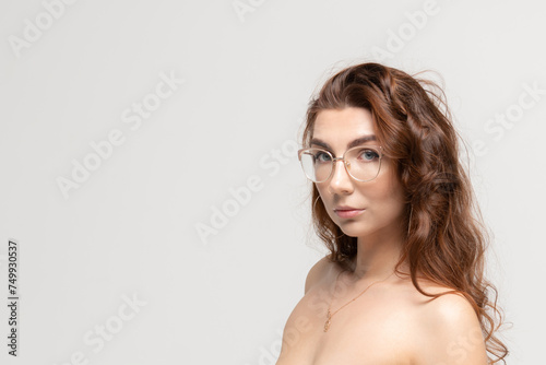 Close-up portrait of beautiful caucasian woman with long brown curly hair and open shoulders wearing eyeglasses against grey background. Soft focus. Copy space for your text. Beauty an skincare theme.