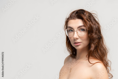 Close-up portrait of beautiful caucasian woman with long brown curly hair and open shoulders wearing eyeglasses against grey background. Soft focus. Copy space for your text. Beauty an skincare theme.