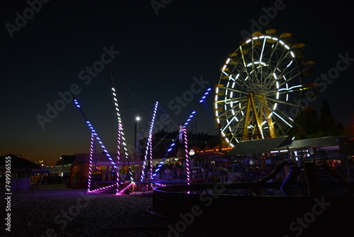 Night entertainment and relaxation on the beach of the Black Sea with attractions  white Ferris wheel  trampoline with a light  blue  yellow color and a children s pool.