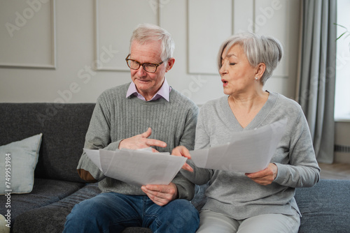 Sad tired disappointed middle aged senior couple sit with paper document. Unhappy older mature man woman reading paper bill managing bank finances calculating taxes planning loan debt pension payment