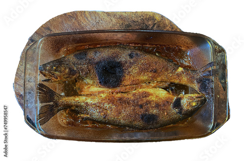 Dwie upieczone ryby, pstrągi w szklanym, żaroodpornym, prostokątnym naczyniu do pieczenia ustawionym na drewnianej desce. Złota, przypieczona skórka. Widok z góry. Przezroczyste tło.