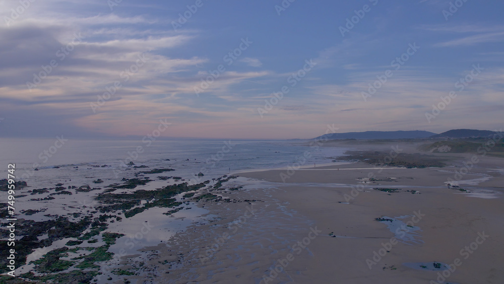 Sea, beach boulders, pebble shore, and waves at sunset. Sea waves breaking on rocks. Beach with sand dunes and dusk sky in Rio de Moinhos, Esposende, Portugal.