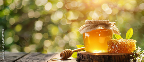 A jar glass full of honey with honeycomb and honey dripper on wooden table, blurred natural green background.