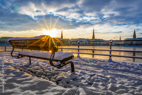 Verschneite Parkbank am Ballindamm in Hamburg morgens im Winter bei Sonnenaufgang, im Hintergrund unscharf die Gebäude an der Alster zu sehen, horizontal