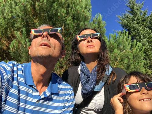 Father, mother and daughter, family viewing solar eclipse with special glasses in a park © jovannig