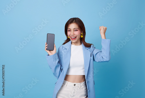 Beautiful Asian teen woman holding smartphone mockup of blank screen and hand up celebration isolated on blue background.