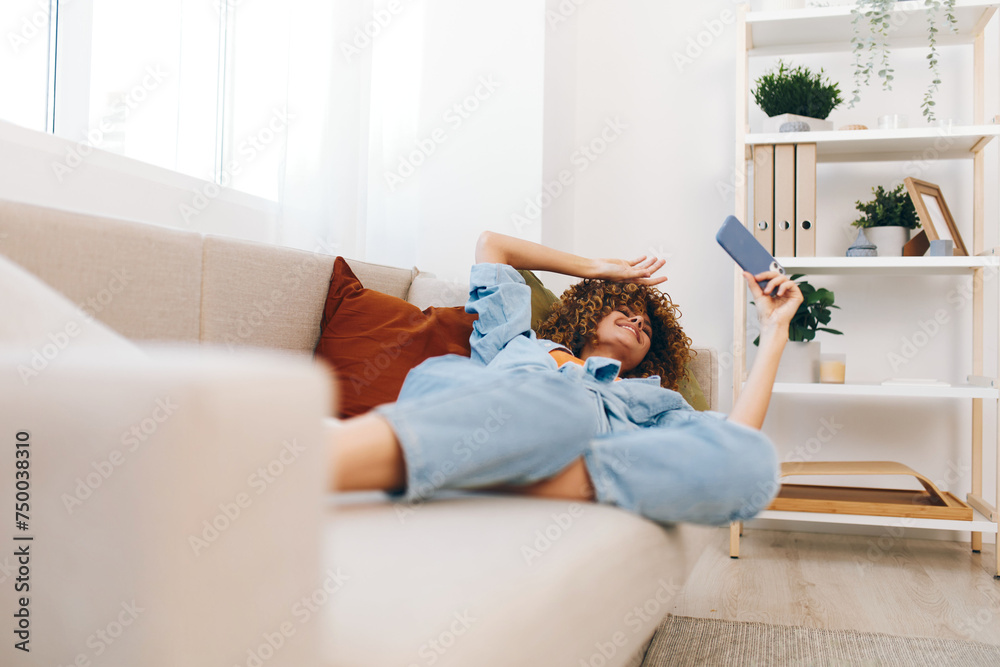 Happy woman sitting on a cozy sofa, holding a mobile phone and smiling while enjoying online games, reading, and taking a selfie at home Happy woman sitting on a cozy sofa, holding a mobile phone and smiling while enjoying online games, reading, and taking a selfie at home
