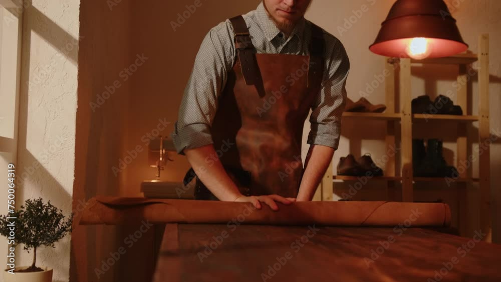 Shoemaker rolls out piece of leather on table in workshop. Artisan works with leather to make handmade shoes.