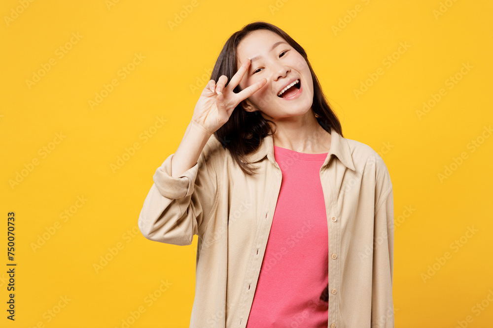 Young smiling happy woman of Asian ethnicity wear pink t-shirt beige shirt pastel casual clothes show cover face eye with victory sign isolated on plain yellow background studio. Lifestyle portrait. Young smiling happy woman of Asian ethnicity wear pink t-shirt beige shirt pastel casual clothes show cover face eye with victory sign isolated on plain yellow background studio. Lifestyle portrait.