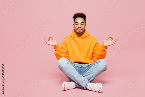 Full body spiritual young man of African American ethnicity wear yellow hoody casual clothes sit hold spread hand in yoga om aum gesture relax meditate try calm down isolated on plain pink background photo