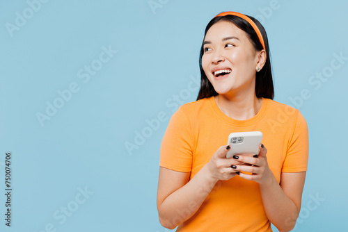Young happy woman of Asian ethnicity wears orange t-shirt casual clothes hold in hand use mobile cell phone look aside on area isolated on plain pastel light blue cyan background. Lifestyle concept.