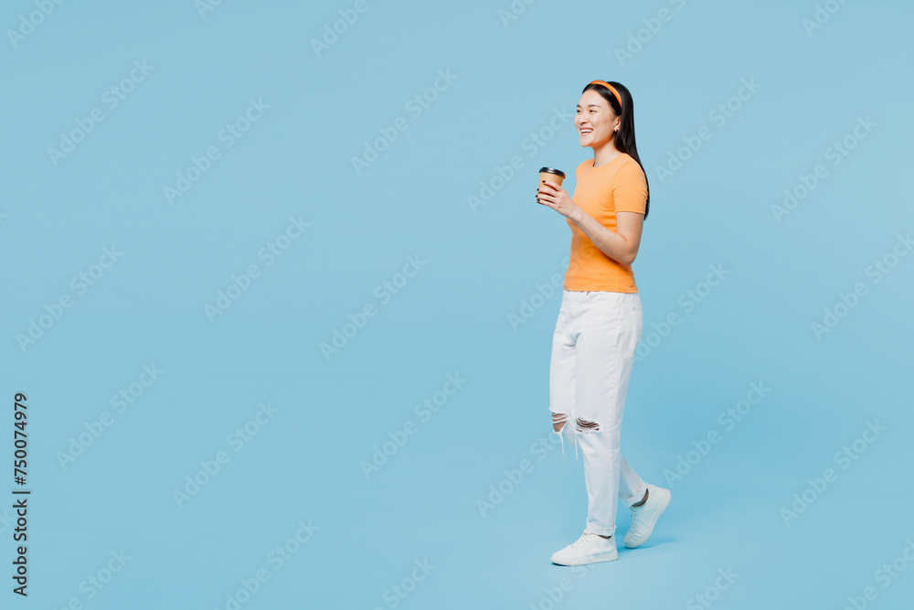 Full body young woman of Asian ethnicity wear orange t-shirt casual clothes hold takeaway delivery craft paper brown cup coffee to go isolated on plain pastel blue cyan background. Lifestyle concept.