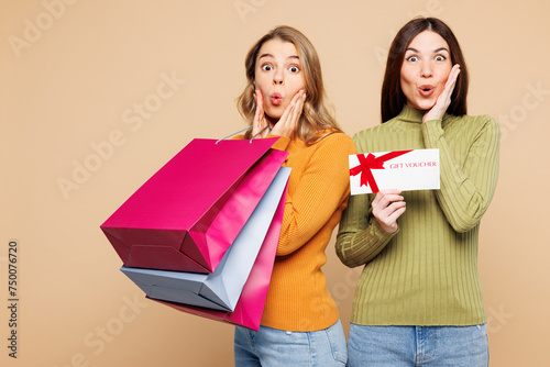 Young amazed friends two women in orange green shirt casual clothes hold shopping package bags gift coupon voucher card for store isolated on plain beige background Black Friday sale buy day concept