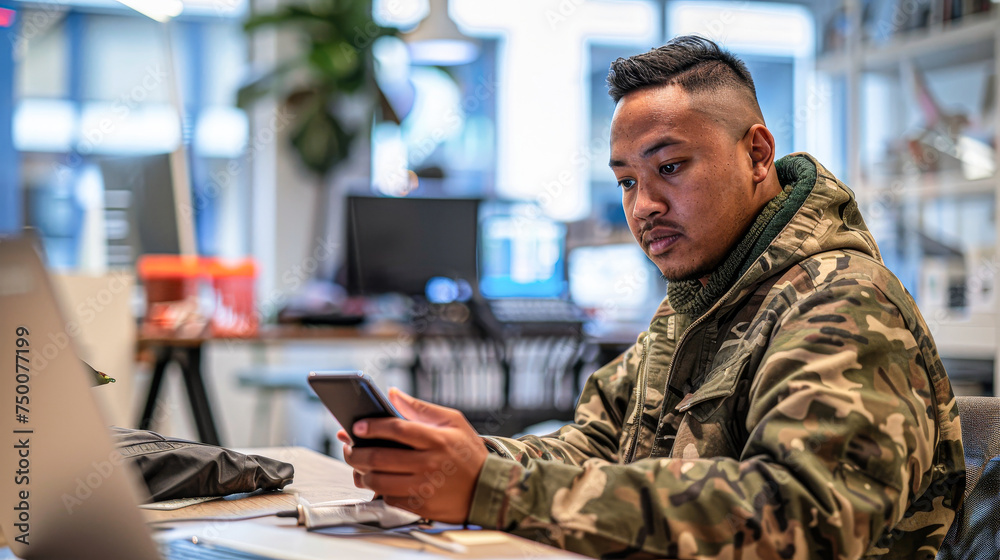 A man in a camo jacket is looking at his cell phone. He is sitting at a desk with a laptop and a potted plant in the background A man in a camo jacket is looking at his cell phone. He is sitting at a desk with a laptop and a potted plant in the background