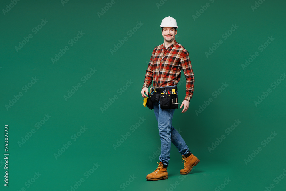 Full body side view young employee laborer handyman man wear red shirt hardhat walk go look camera isolated on plain green background. Instruments for renovation apartment room. Repair home concept. Full body side view young employee laborer handyman man wear red shirt hardhat walk go look camera isolated on plain green background. Instruments for renovation apartment room. Repair home concept.