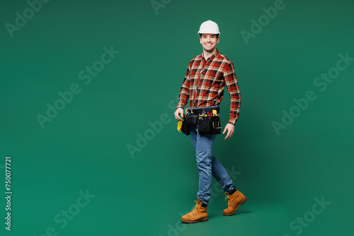 Full body side view young employee laborer handyman man wear red shirt hardhat walk go look camera isolated on plain green background. Instruments for renovation apartment room. Repair home concept. © ViDi Studio