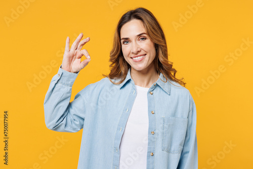 Young smiling happy cheerful woman she wear blue shirt white t-shirt casual clothes showing okay ok gesture looking camera isolated on plain yellow wall background studio portrait. Lifestyle concept.