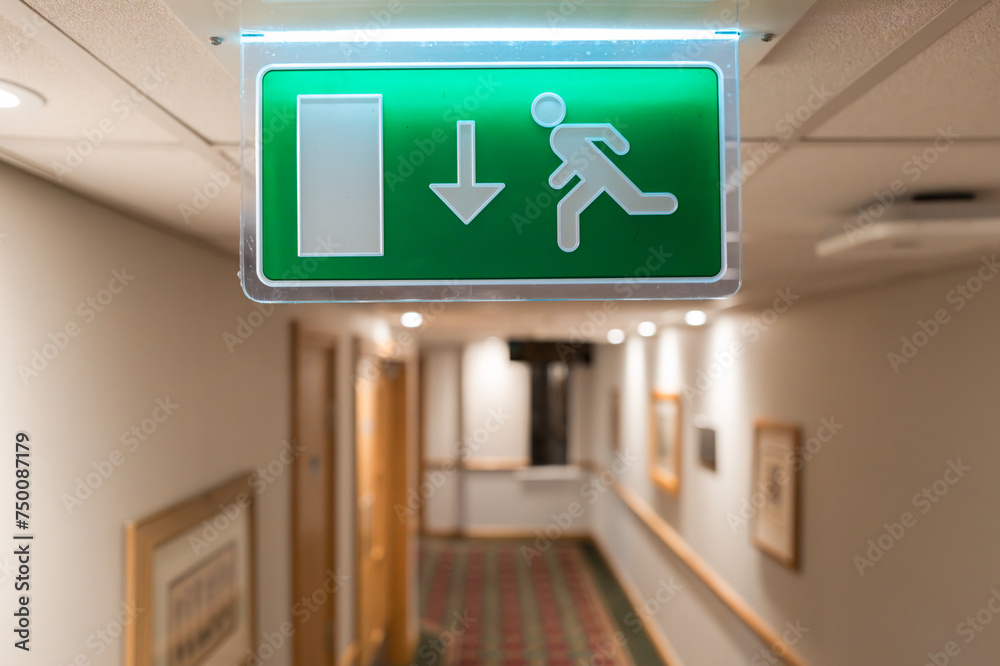 Shallow focus of a generic Fire Escape sign seen hanging from the ceiling of a hotel lobby in the UK. The hotel corridor shows some of the room doors and wall paintings. Shallow focus of a generic Fire Escape sign seen hanging from the ceiling of a hotel lobby in the UK. The hotel corridor shows some of the room doors and wall paintings.