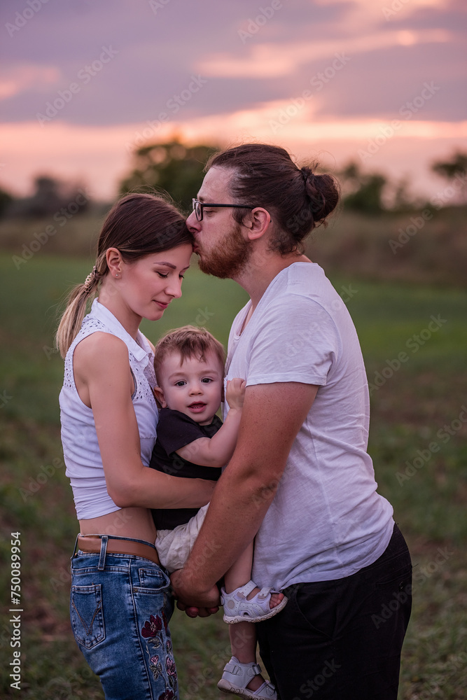 Diversity stylish father kisses mothers forehead while holding little son, concept of beautiful family connection against vibrant sunset. Family ties, togetherness. Close up portrait