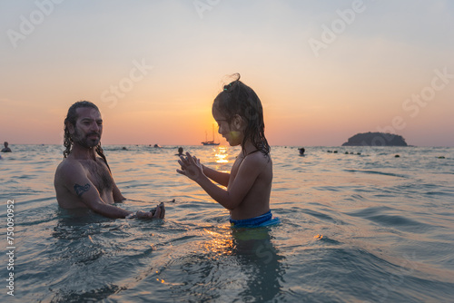 A father carries his child to play in sea water by the beach at sunset. .The father is taking the opportunity to bond with his child and enjoy a peaceful evening. .The sun is on the palm of his hand.