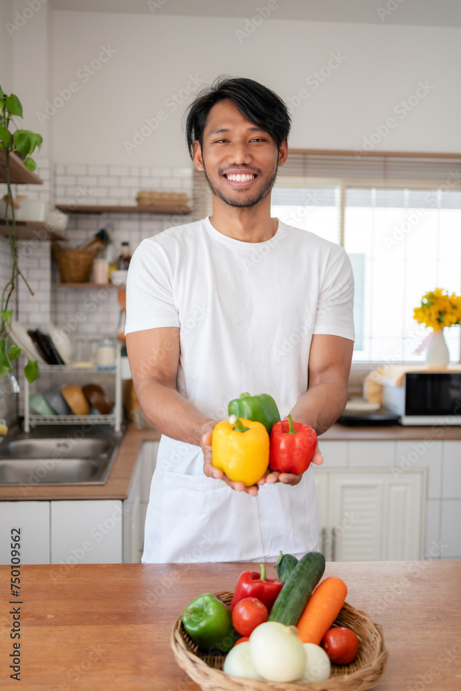 Happy asian male chef holding colorful bell peppers and standing in a home kitchen surrounded by fresh vegetables. Happy asian male chef holding colorful bell peppers and standing in a home kitchen surrounded by fresh vegetables.