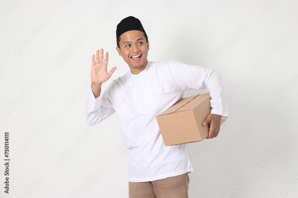 Portrait of excited Asian muslim man in koko shirt with peci carrying cardboard box while saying hi, greeting to someone. Going home for Eid Mubarak. Isolated image on white background