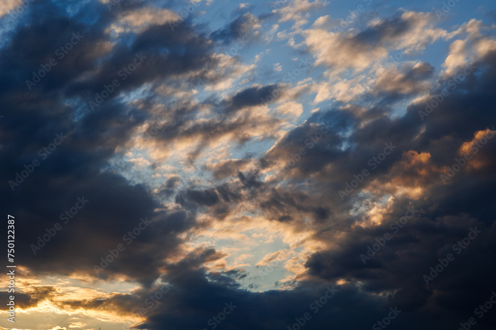 visuale in primo piano di un cielo azzurro, al tramonto, parzialmente nuvoloso, con nuvole color grigio e blu scuro, illuminato lateralmente dalla luce arancione del sole