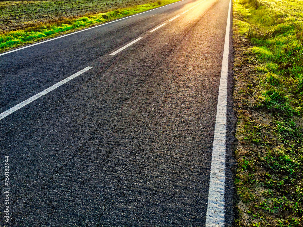 Sunlight casts shadows on an empty road surrounded by grass. Sunlight casts shadows on an empty road surrounded by grass.