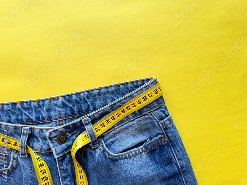 Close up of blue denim jeans with yellow measuring tape belt on vivid yellow background with copy space. Fashion, dieting, and body measurements. Top view.