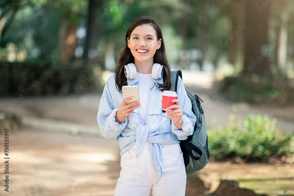 A cheerful student with a warm smile enjoys a walk in the park, holding a smartphone