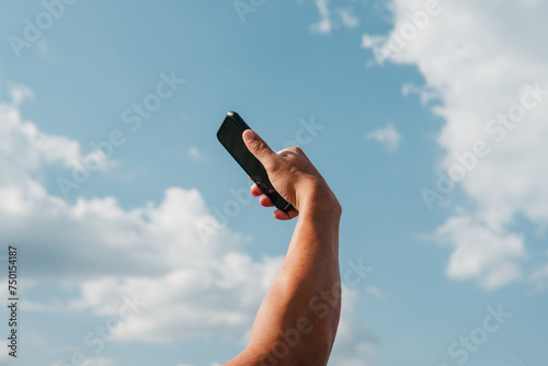 Photography A man's arm extends upwards, holding a smartphone against a backdrop of fluffy c