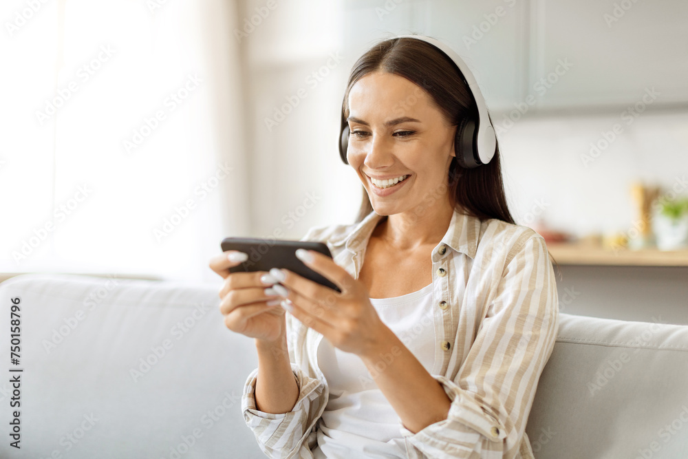 Smiling young woman wearing wireless headphones using her smartphone at home Smiling young woman wearing wireless headphones using her smartphone at home