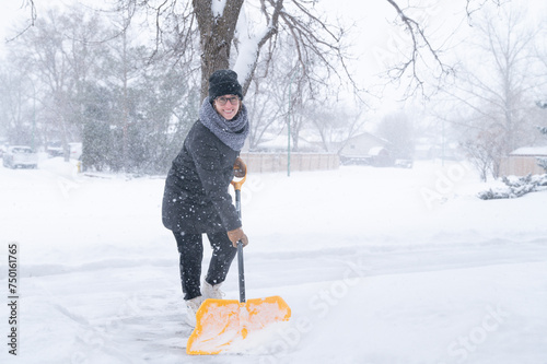 A young woman dressed in a winter coat, gloves, hat and scarf holding a yellow snow shovel, clearing a pathway after a blizzard