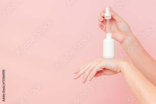 Hands holding glass bottle with dropper lid. White container with cosmetic product, serum (essential oil ) on pink background. Concept of beauty
