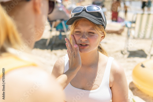 Against the backdrop of the summer beach, a mother tenderly applies sunscreen to her daughter's face as they sit in their swimsuits, embodying the essence of seaside relaxation and parental care.