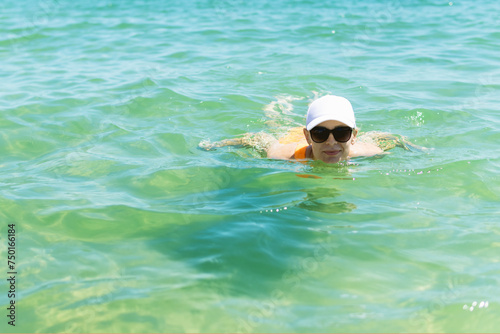 A happy woman in a swimsuit, sunglasses, and white cap enjoys a swim in the sea during summer, capturing the essence of carefree oceanic bliss. © PopOff