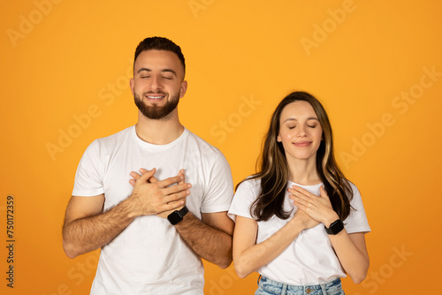 Grateful young couple with eyes closed and hands on hearts, expressing sincere emotions and appreciation