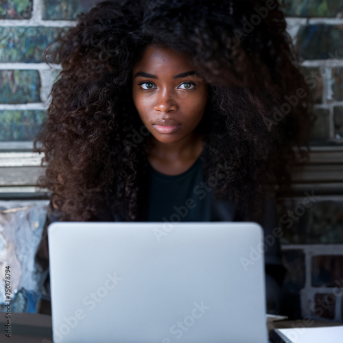 A young woman with voluminous curly hair sits in front of a laptop in a café setting. Her intense gaze is directed towards the camera, giving the impression of focus and determination.