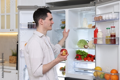 Happy man holding apple near refrigerator in kitchen