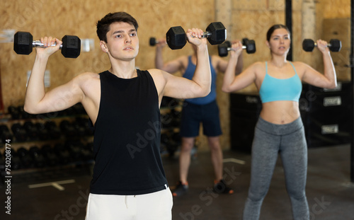 Young dedicated man doing exercises with dumbbells near other people in gym