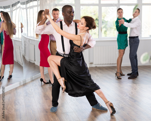 Woman in elegant evening dress dances incendiary tango with African American male partner during choreography lesson. Group training and rehearsal, preparation for competitions