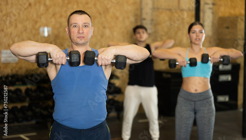 Sportive strong middle-aged male in activewear lifting dumbbells during group workout class in gym indoors. Functional training concept © JackF