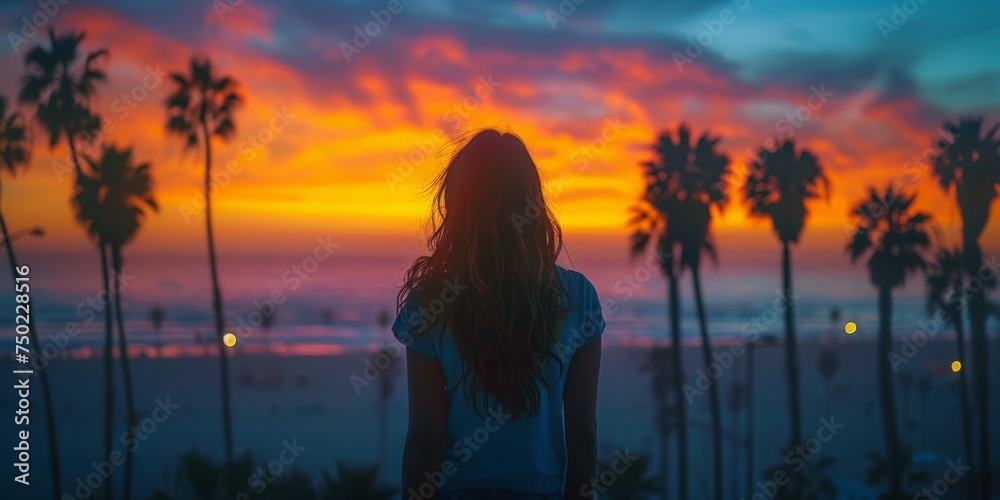 A person is standing and looking at the sunset over the ocean surrounded by palm trees. The sky is full of colors ranging from orange to purple - obrazy, fototapety, plakaty 