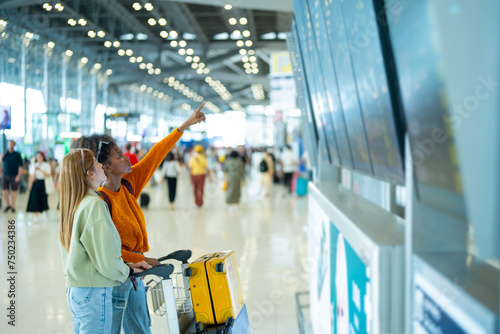 Woman friends looking flight schedule on timetable and go to check-in and boarding gate together in airport terminal. Attractive girl traveler travel by airplane transportation on holiday vacation.