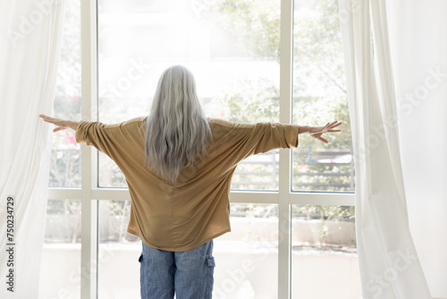 Back view of senior woman with long naturally grey hair in casual opening drapes, parting white transparent curtains, veils at large window, enjoying view of terrace view photo