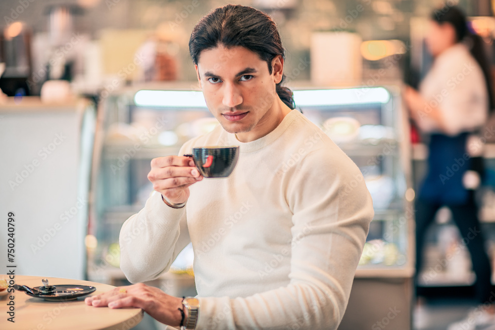 A brunette Hispanic man with long hair drinking coffee from a black mug.