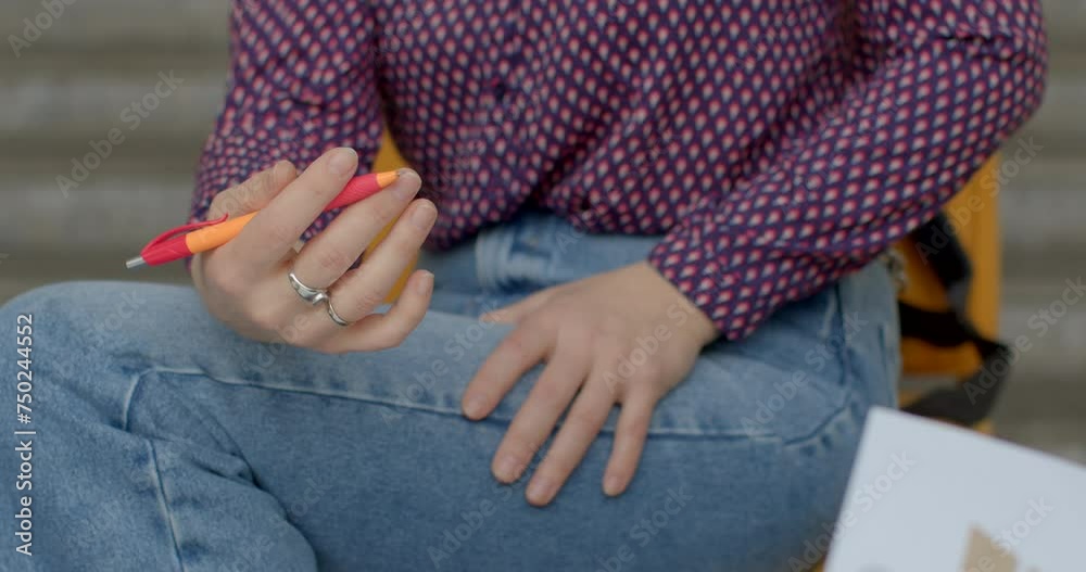 Educational Process: Female Student or teacher Casual Style in Taking Notes. Student's hands holding a pen and making notes during an educational process. Orange color pen in hand, blue jeans. Educational Process: Female Student or teacher Casual Style in Taking Notes. Student's hands holding a pen and making notes during an educational process. Orange color pen in hand, blue jeans.