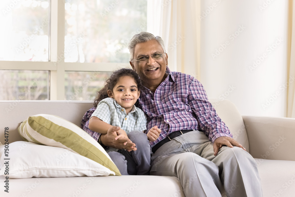 Happy caring older Indian grandpa and cute little granddaughter kid hugging with love, affection, sitting on home couch, looking at camera, smiling for family portrait, laughing