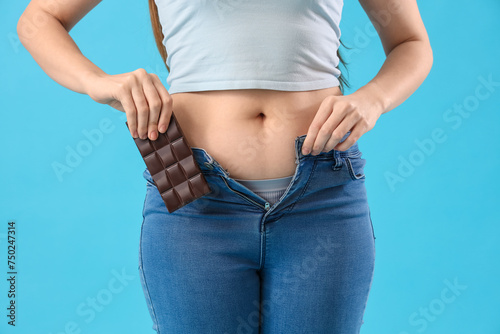 Young woman in tight jeans with chocolate on blue background, closeup. Weight gain concept